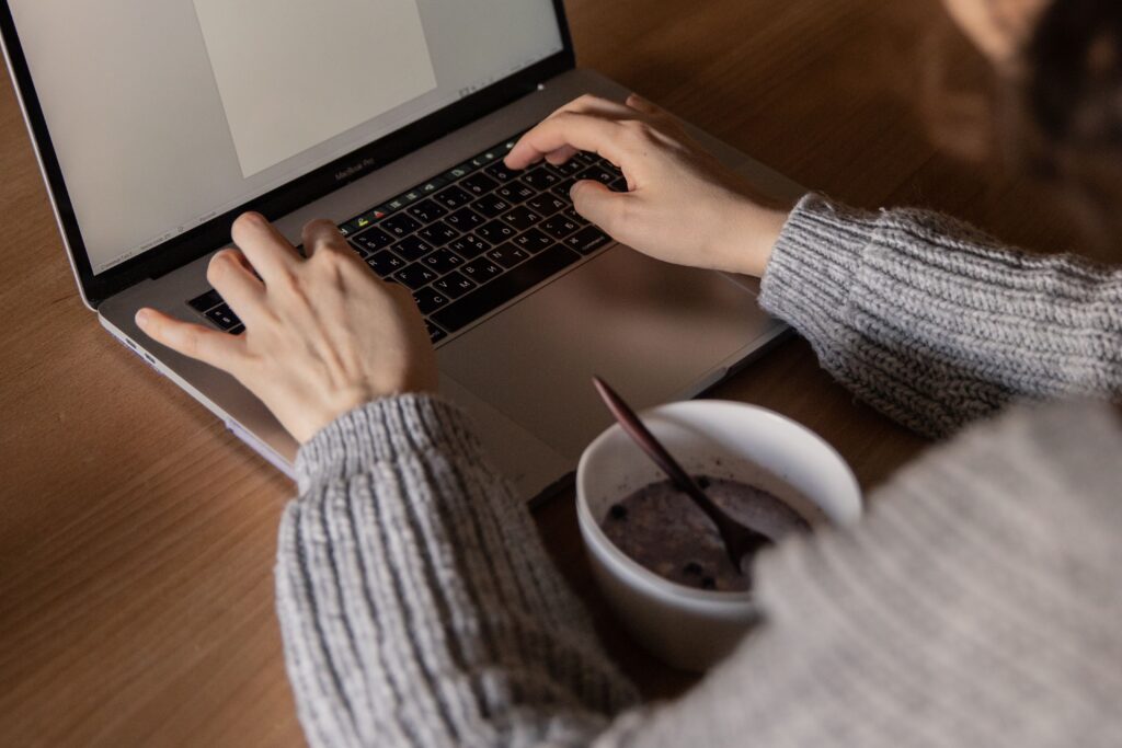 From above unrecognizable woman in gray sweater sitting at wooden table with food in bowl and creating document while working on remote project at home